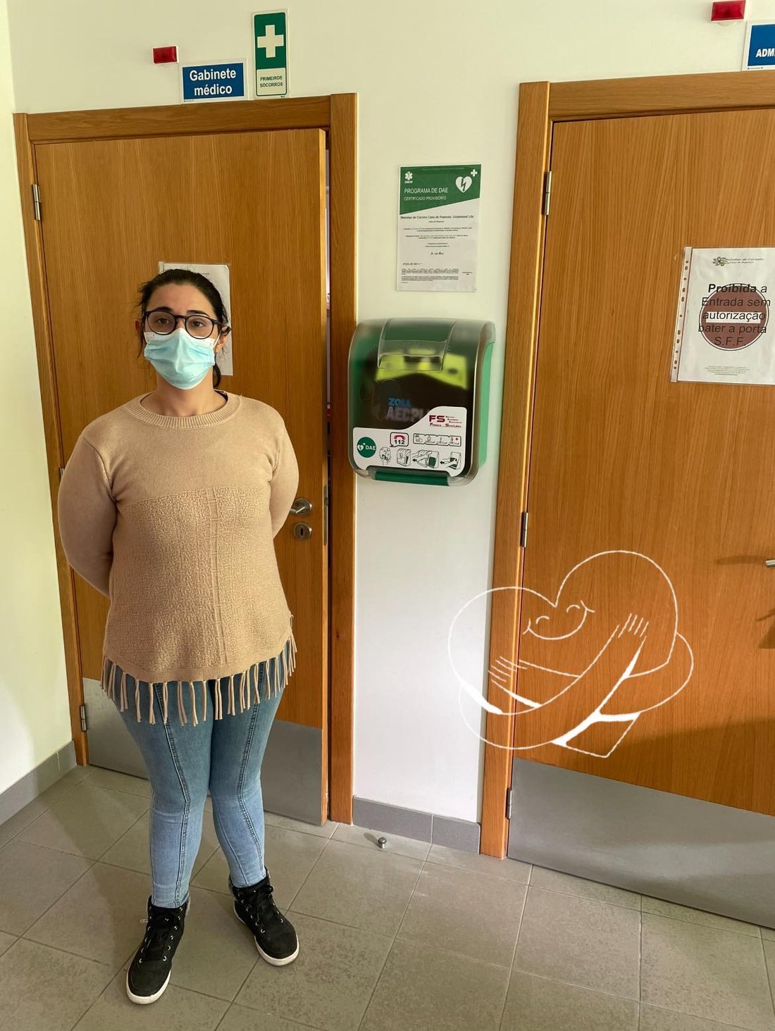 Colaboradora junto ao armário DAE instalado num corredor, com certificado do Programa DAE afixado na parede